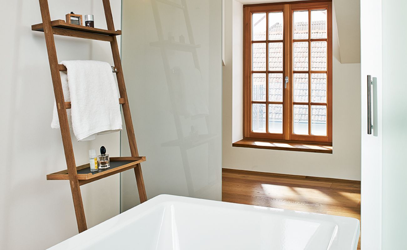 Moderne Badezimmeransicht mit freistehender Badewanne, Holzregal und großen Fenstern, die Tageslicht hereinlassen.